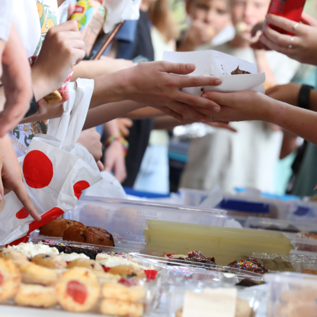 Hands exchanging baked goods at a crowded vendor table, with cookies, pastries, and desserts in view.