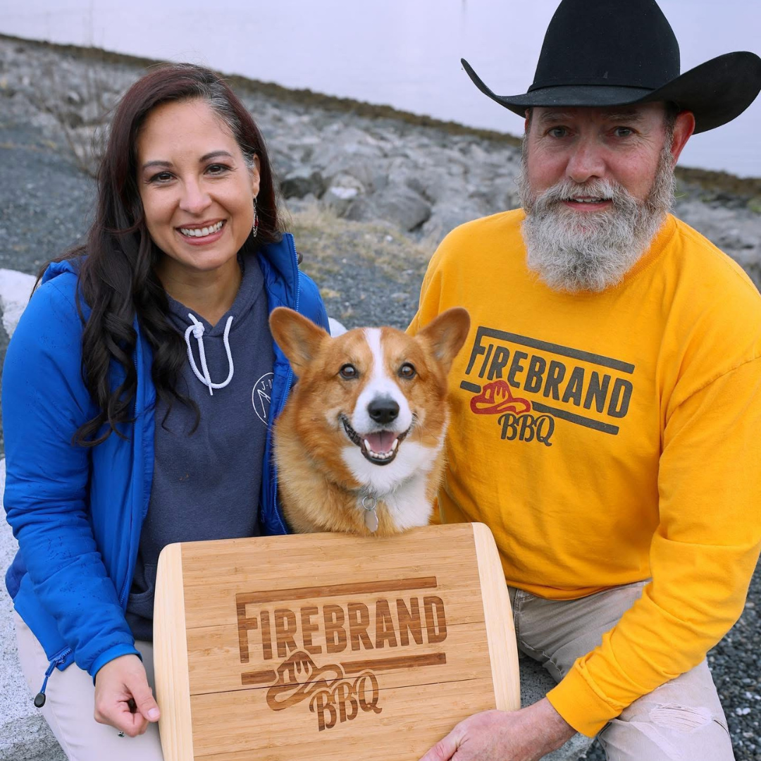 Owners of Firebrand BBQ pose with their corgi and a branded cutting board near the Alaska waterfront.