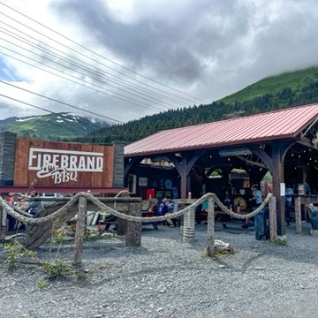 Exterior view of Firebrand BBQ restaurant in Seward, Alaska with mountain scenery and outdoor seating area.
