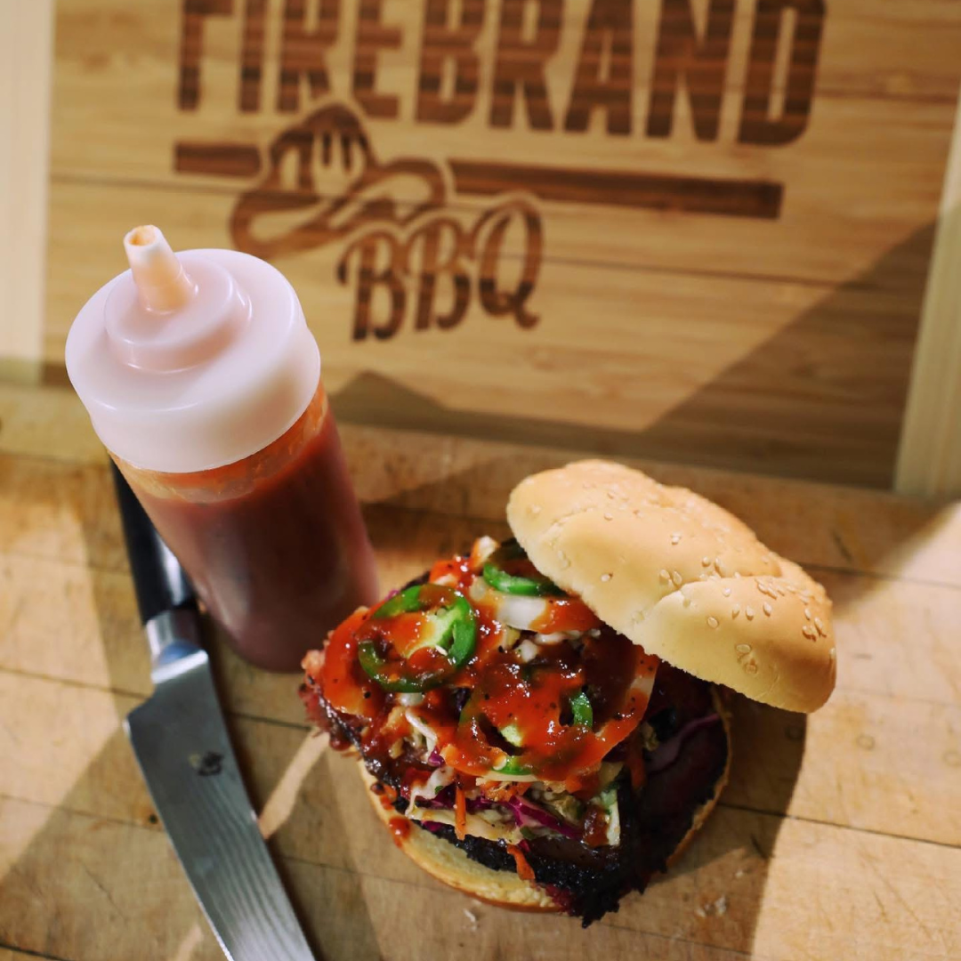 Smoked barbecue sandwich from Firebrand BBQ topped with sauce and vegetables on a cutting board beside a bottle of barbecue sauce.