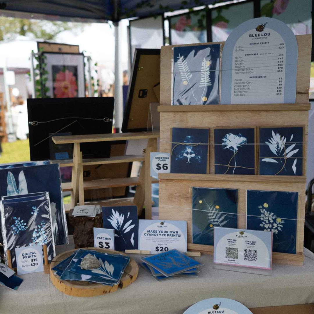 Blue Lou Art market booth with cyanotype prints, cards, and small handmade items arranged on a wooden display.
