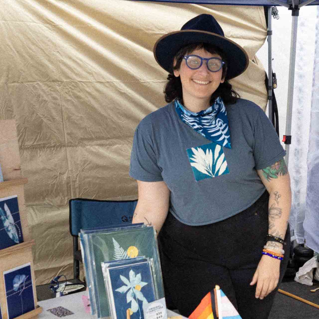Artist standing behind a booth displaying Blue Lou cyanotype prints, cards, and handmade goods.