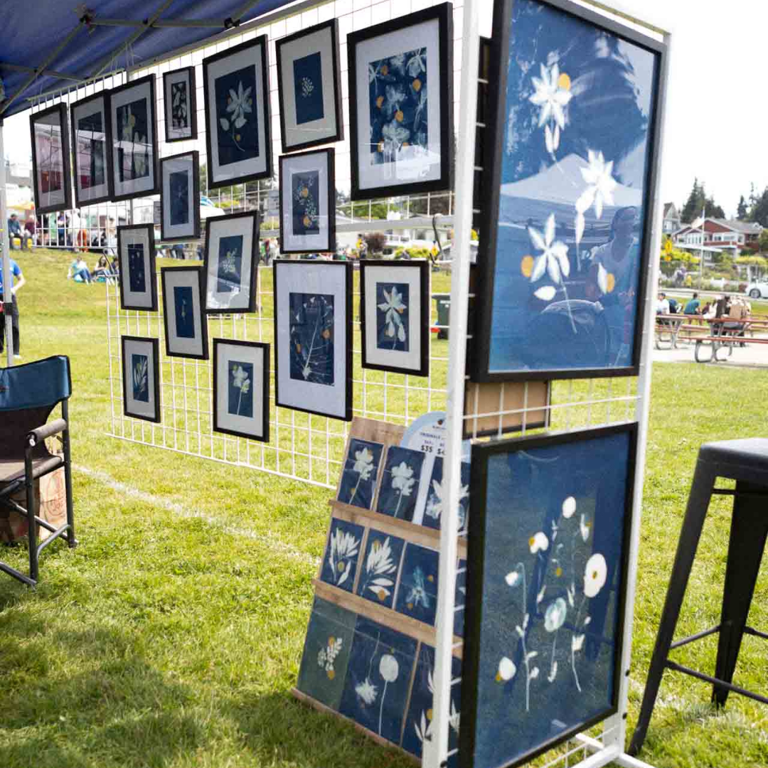 Outdoor booth displaying framed botanical prints and blue cyanotype artwork at a community market.