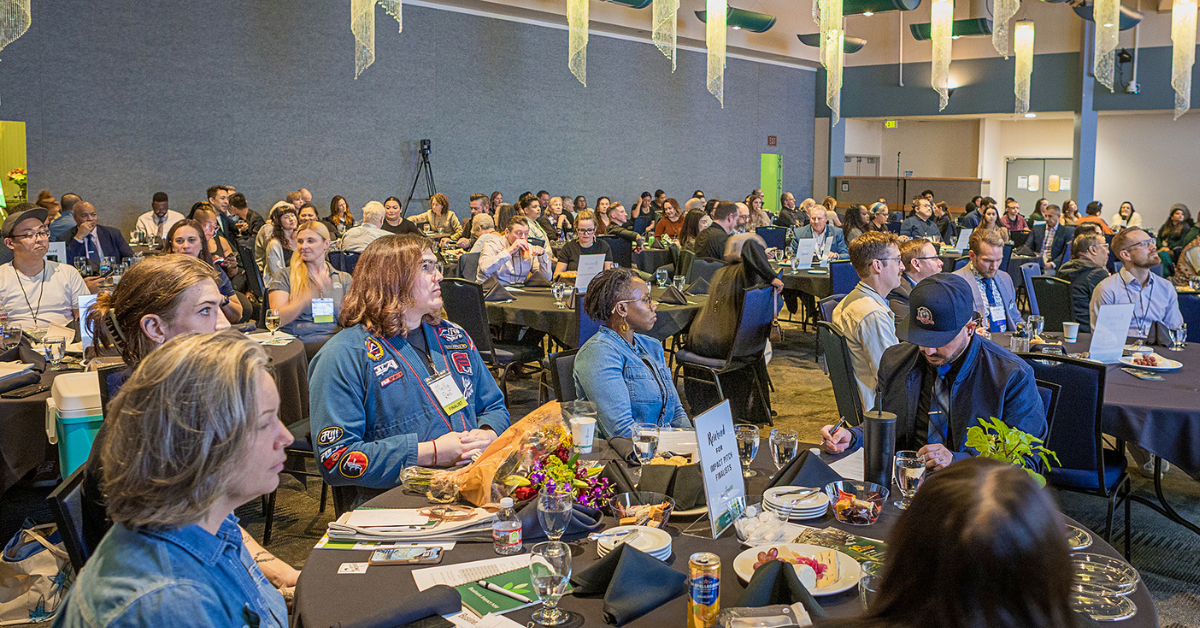 Audience members seated at banquet tables watch the IMPACT Pitch program in a large event space.