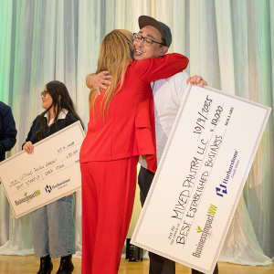 A Mixed Pantry LLC hugs a judge onstage while holding a $10,000 Best Established Business award check at IMPACT Pitch.